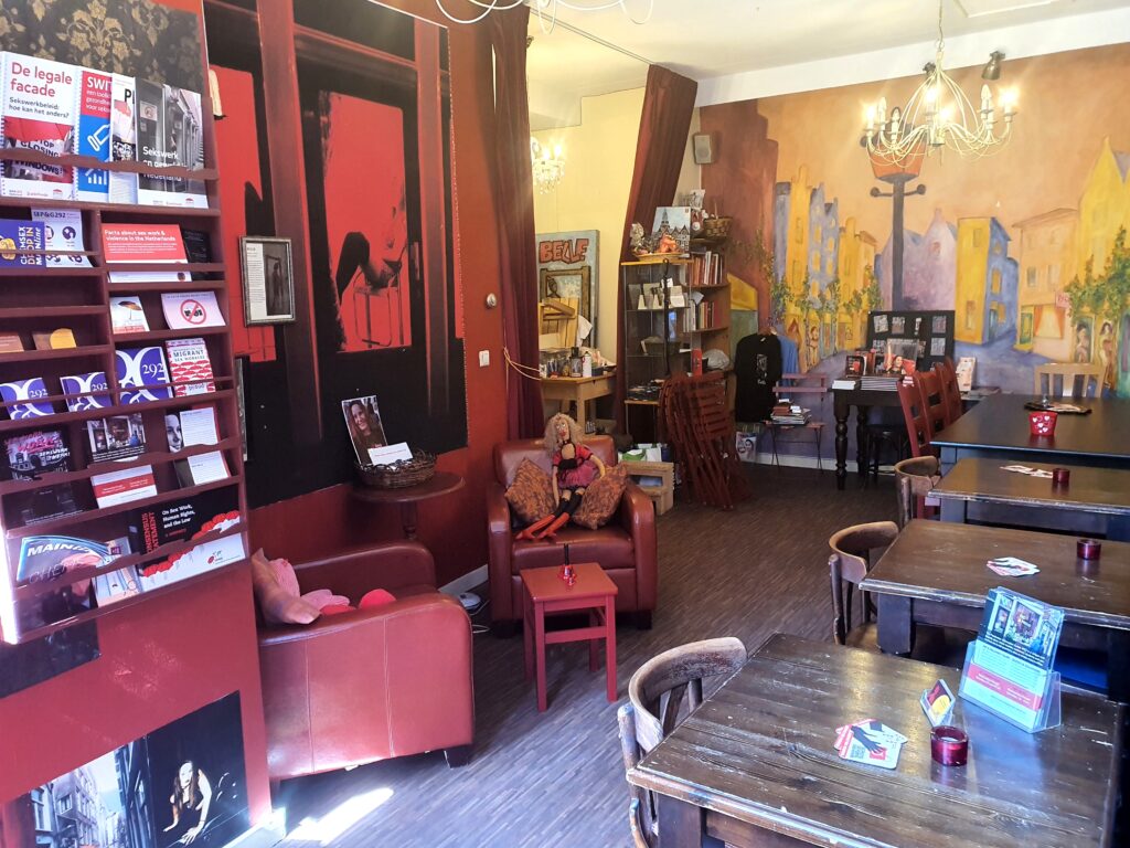 A picture of the interior of PIC, which contains two red chairs and 4 wooden tables. To the left are shelves with pamphlets and the back wall is covered with a painting of the Red Light District.